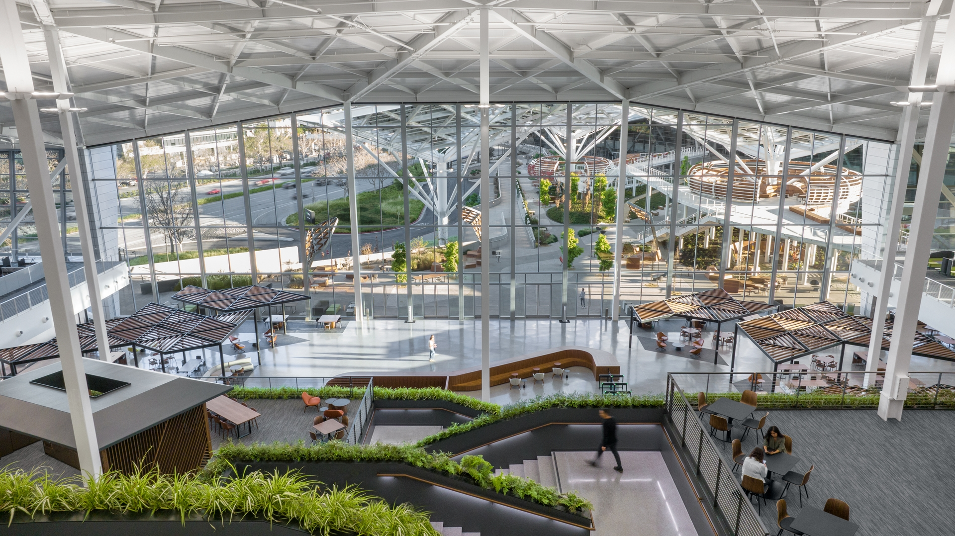 Aerial interior view of NVIDIA's Voyager building atrium, showing a glass-walled multi-level workspace with wood-slat canopy seating areas, greenery, and an open staircase connecting floors.