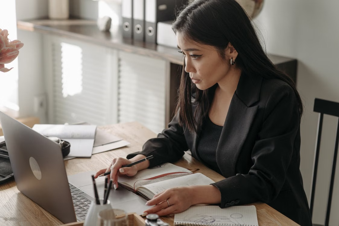 A woman in a black blazer sits at a wooden desk, focused on her laptop while writing in an open notebook, with office supplies and folders in the background.