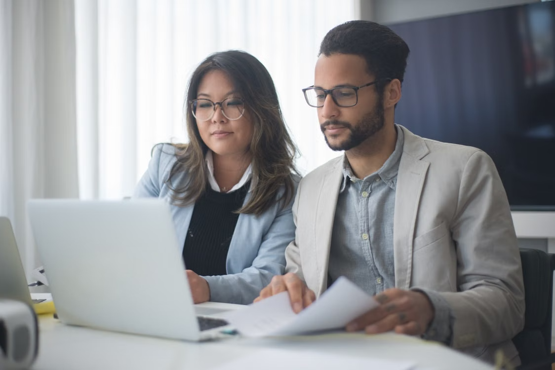 Two colleagues in a bright office reviewing documents together at a laptop, focused on a discussion.