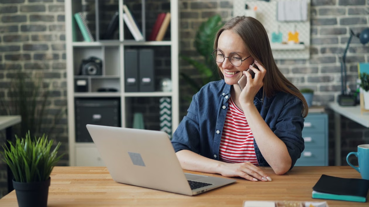 Person smiling while talking on the phone and working on a laptop at a desk in a modern home office.