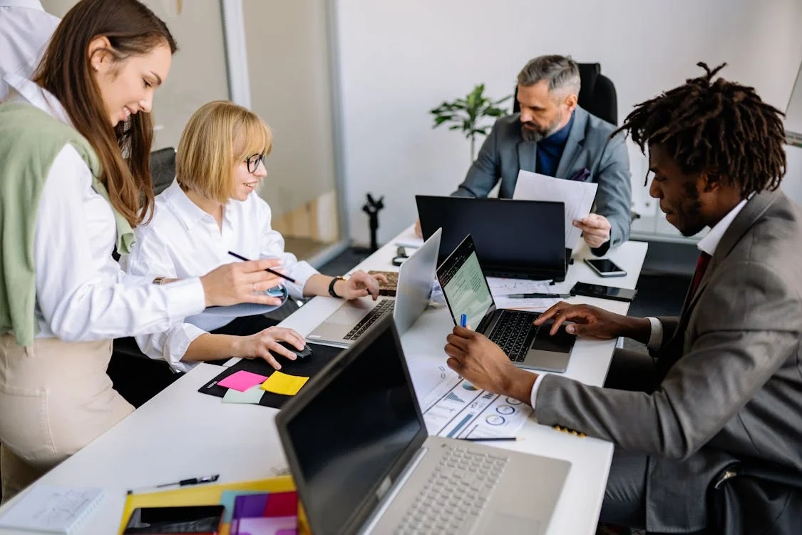 Four professionals review documents and work on laptops around a conference table, illustrating collaborative oversight and decision-making for AI governance.
