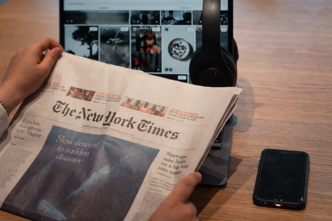 Person reads a printed copy of The New York Times beside a tablet displaying digital images, headphones, and a smartphone, illustrating the intersection of publishing content and digital media.