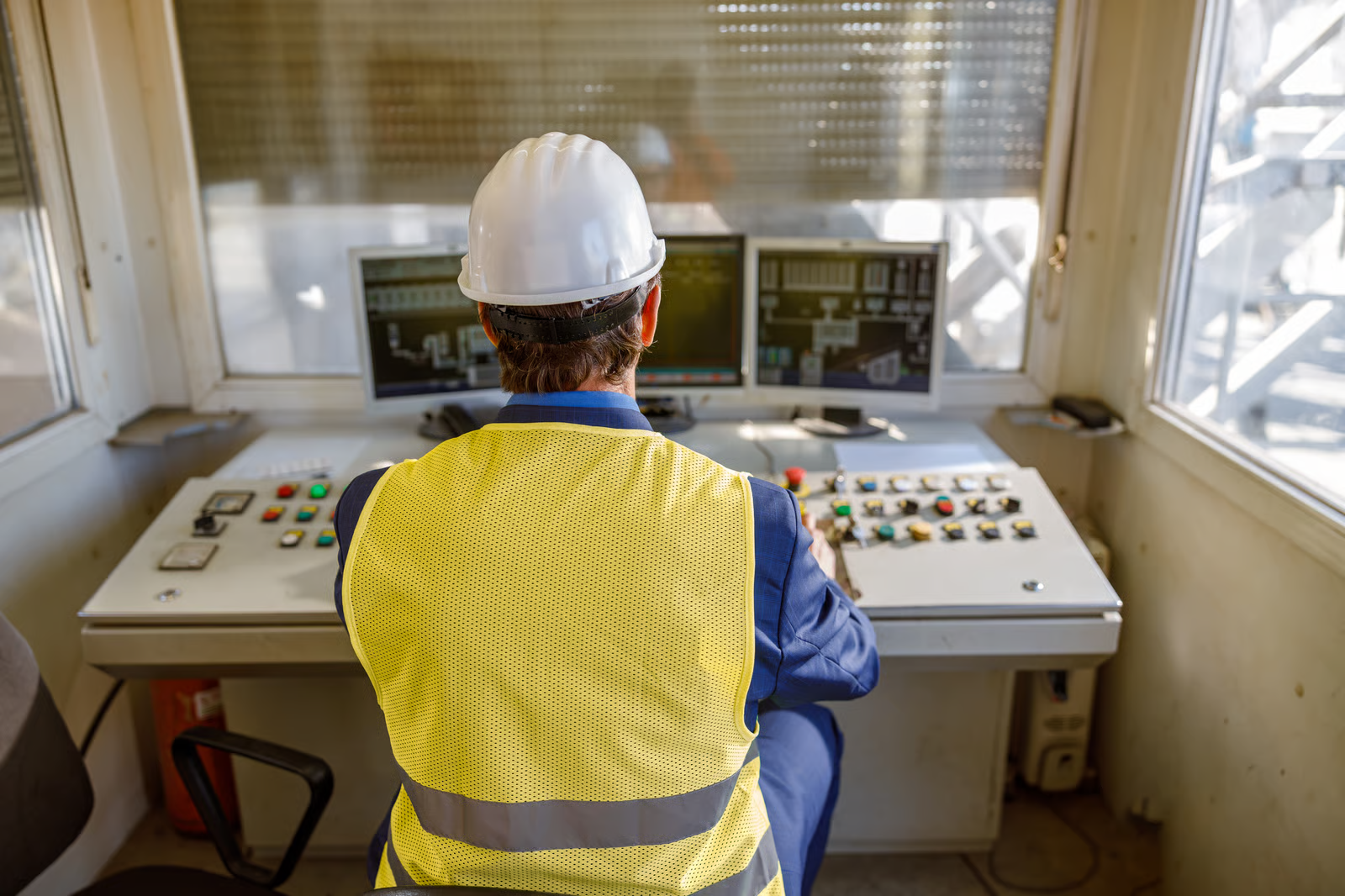 Industrial worker in a hard hat and high-visibility vest monitors equipment from a control room, illustrating AI-supported industrial operations and predictive maintenance.