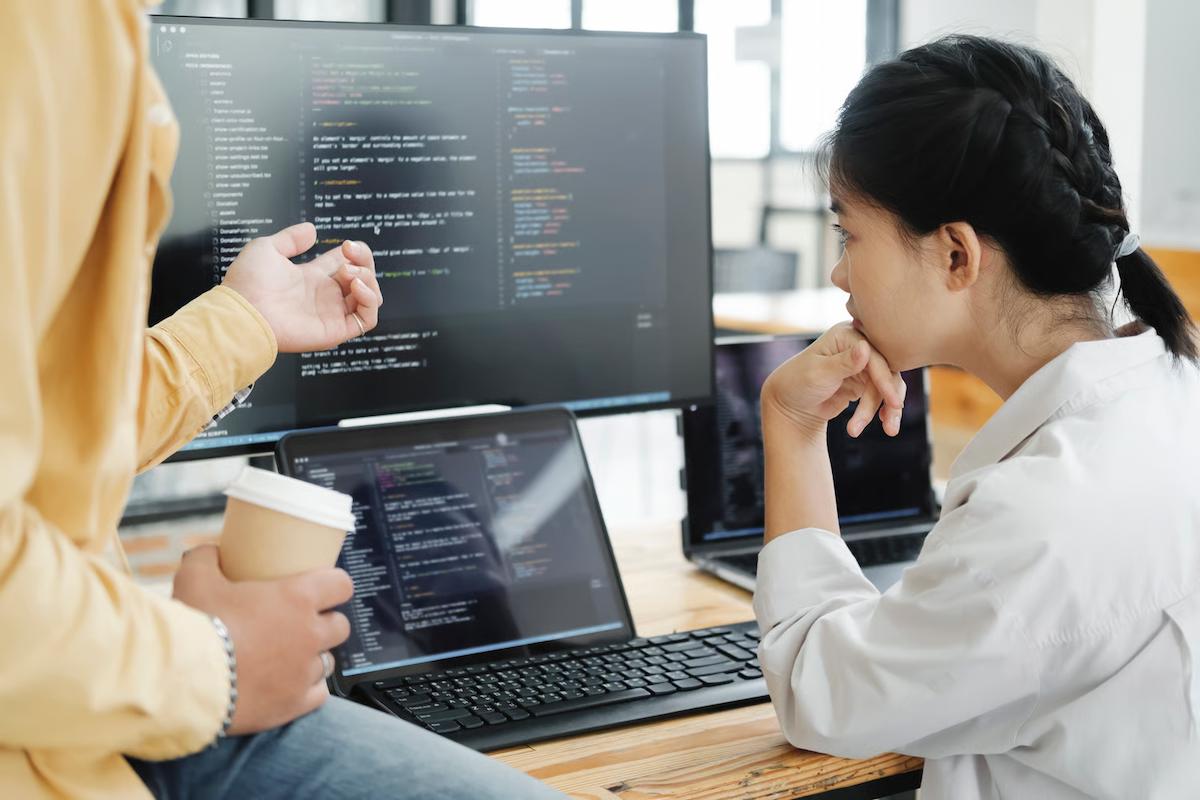 Two software developers reviewing code on a large monitor and laptop, illustrating an enterprise code review comparison of Claude Opus 5.5 vs Opus 5