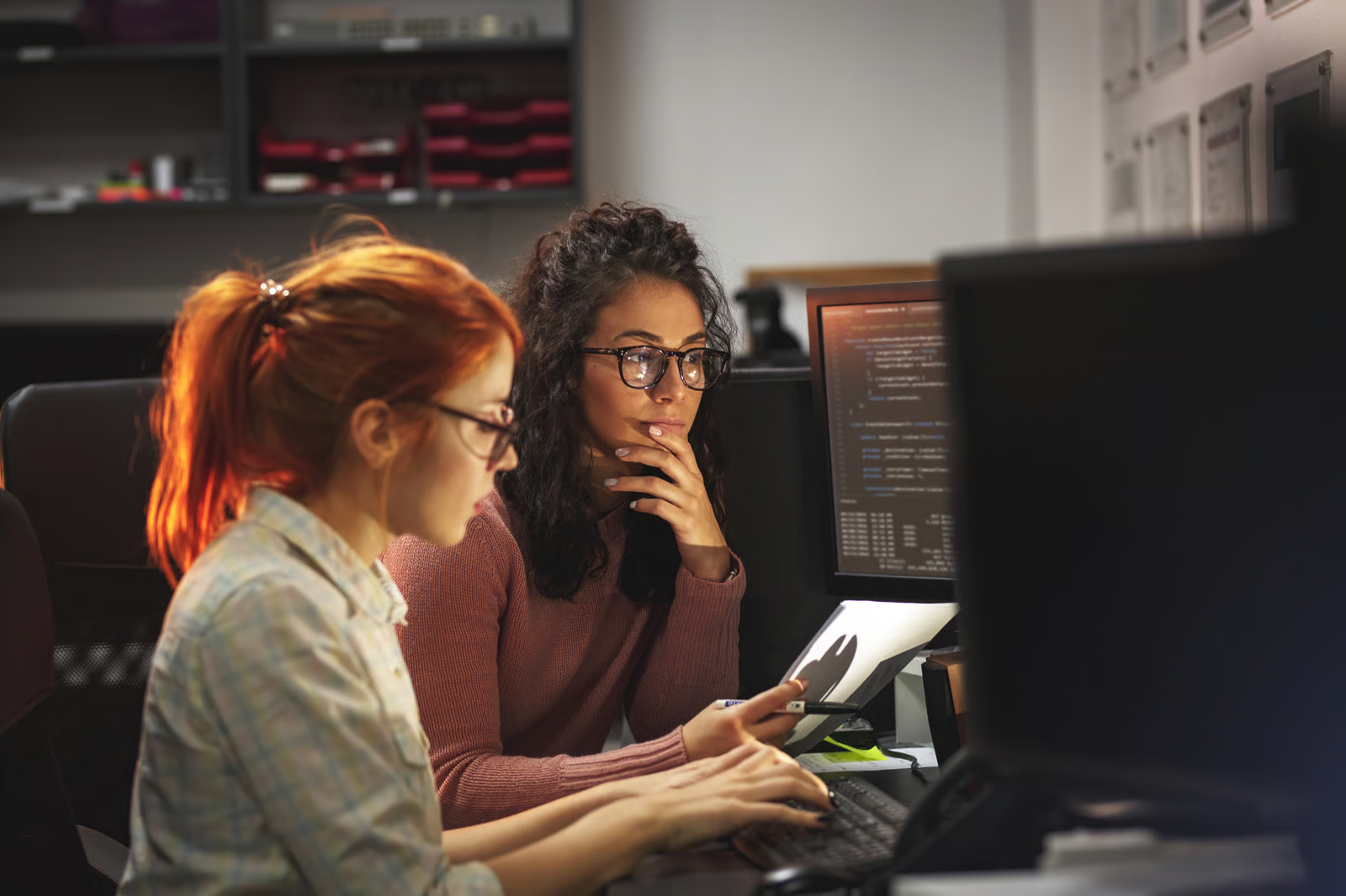 Two developers reviewing code on a monitor while one types and the other checks printed notes, illustrating teams planning a migration from Claude Opus 5 to Opus 5.5