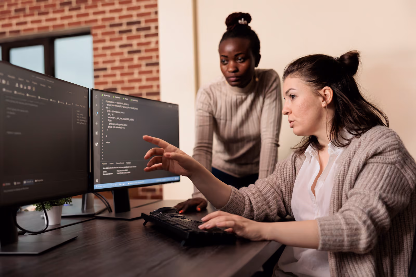 Developer pointing at code on dual monitors while explaining it to a colleague, illustrating teams reviewing Gemini, Claude, and ChatGPT benchmark results