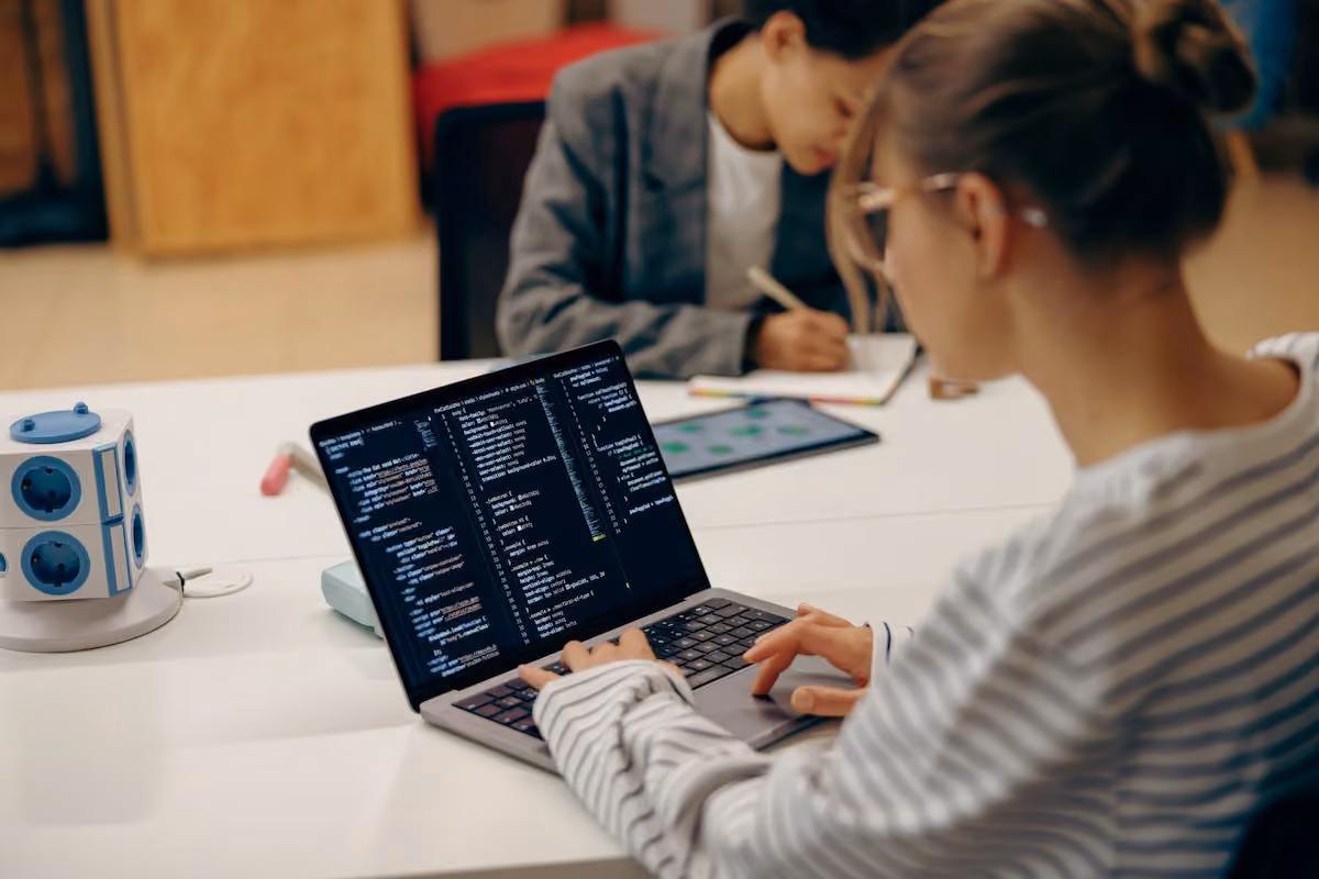 Developer writing code on a laptop while a colleague takes notes in the background, illustrating teams upgrading from Claude Sonnet 5 to Claude Sonnet 5.5 for coding work