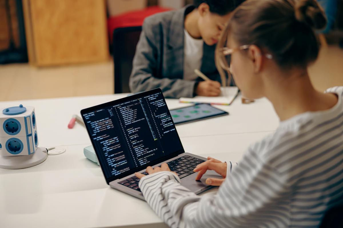 Developer writing code on a laptop while a colleague takes notes in the background, illustrating teams upgrading from Claude Sonnet 5 to Claude Sonnet 5.5 for coding work
