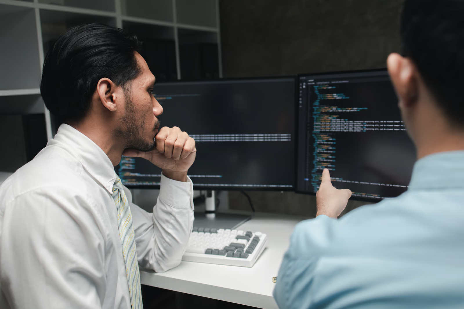 Two developers reviewing code on dual monitors as one points to a line on screen, illustrating teams checking breaking changes before migrating from Claude Sonnet 5 to Sonnet 5.5