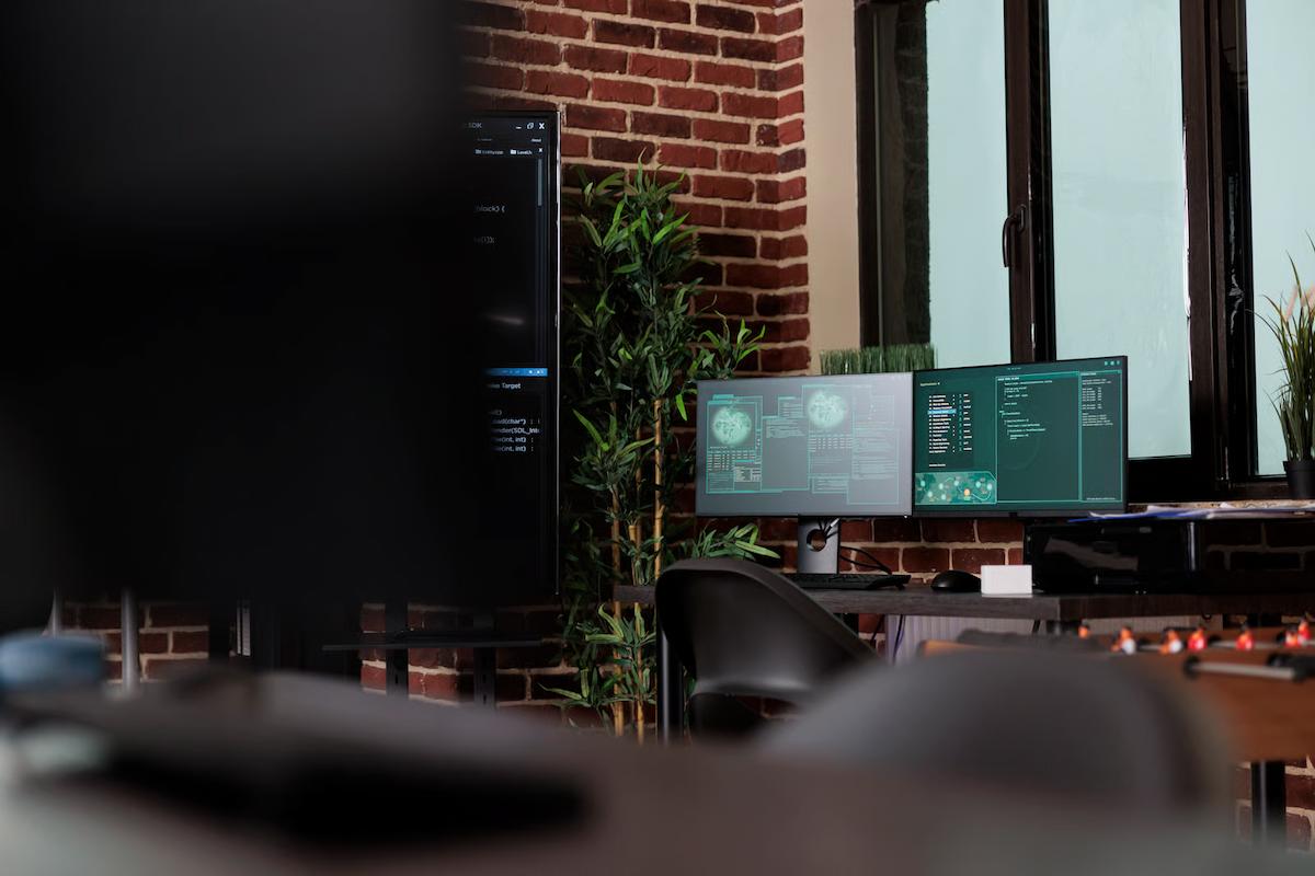 An empty office with a red brick wall, a tall plant, and a desk with dual monitors showing data windows and code, beside a large vertical screen of code, illustrating the data platform and engineering work behind a semantic layer for AI applications