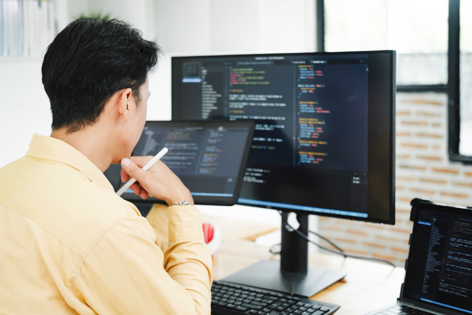 A developer in a yellow shirt holds a stylus to his chin while reviewing code on a desktop monitor, with a tablet and laptop also showing code, illustrating an engineer working through how a solution should run before building it into production