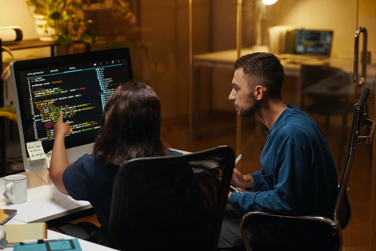 Two developers sit at a desk in a warmly lit office as one points to JavaScript code on a desktop monitor and the other leans in to follow along, illustrating teams testing Claude Sonnet 5.5 and GPT-6.1 Sol on agentic coding work