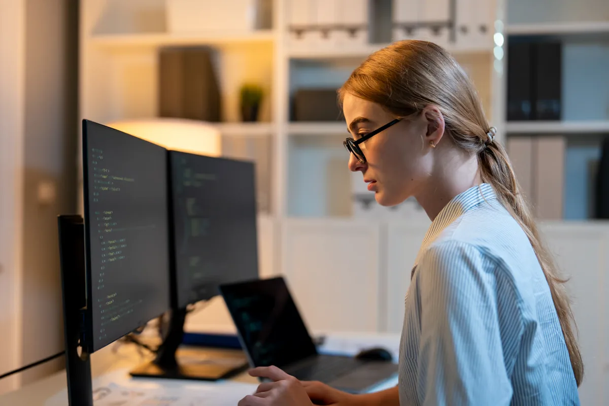 A developer in glasses and a striped shirt works on a laptop in front of dual monitors showing code in a softly lit office, illustrating teams running Claude Sonnet 5.5 and GPT-6.1 Sol on the same tasks to compare accuracy, speed, and cost