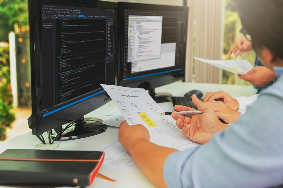 An engineer points a pen at a highlighted section of a printed database development diagram beside dual monitors showing code, illustrating a team checking AI-generated code changes against engineering standards when evaluating coding LLMs