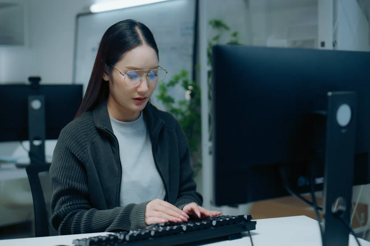 An engineer in glasses types at a desktop computer in a dimly lit office with a whiteboard behind her, illustrating a forward-deployed engineer working in sync with a customer team’s hours to resolve questions the same day