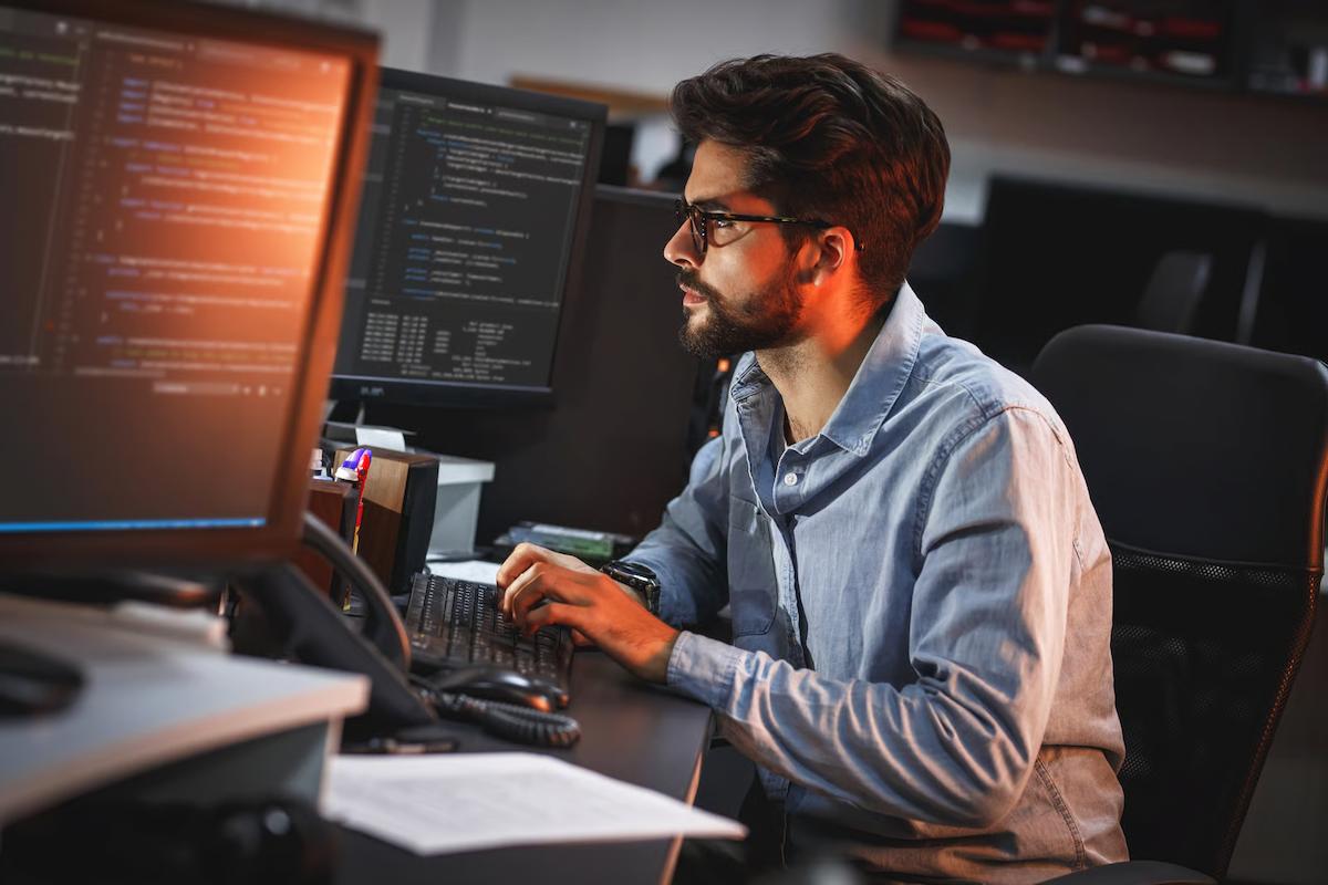 An engineer in glasses and a denim shirt types at a desk with several monitors showing code and terminal output, illustrating the production code and technical skills that define a strong forward-deployed engineer
