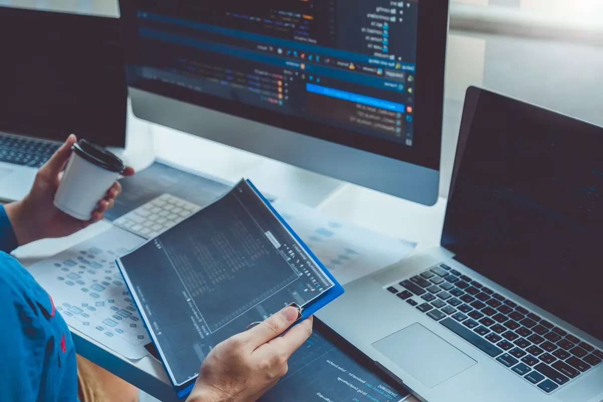 A developer holding a coffee reviews a printed code report beside a monitor, laptop, and a flowchart on the desk, illustrating a forward-deployed engineer mapping a business process and framing the problem before building an AI system