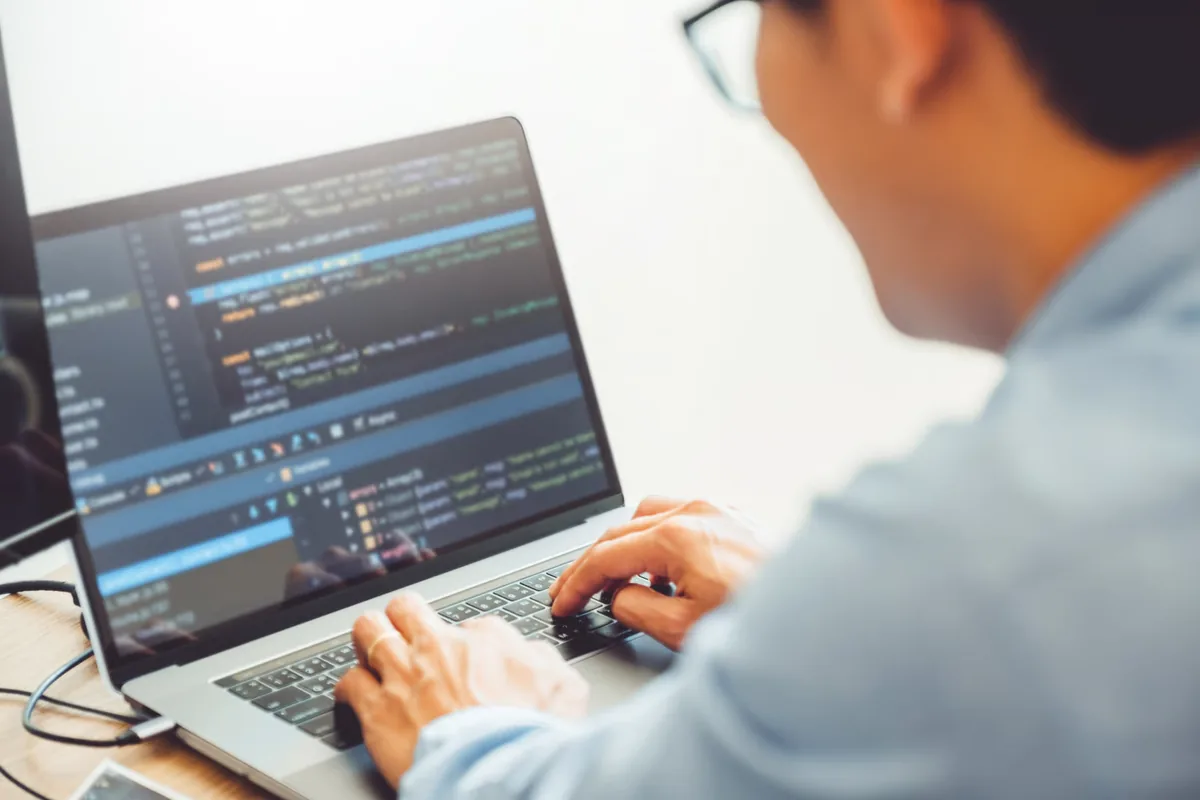 A developer in glasses types on a laptop showing code in an editor with a breakpoint set and a debugging panel open, illustrating a forward-deployed engineer testing and debugging an AI system before people rely on it