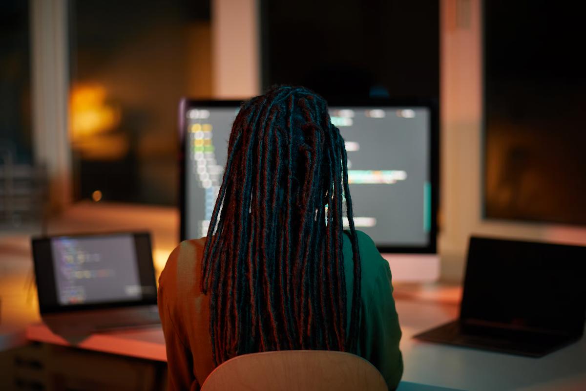 A developer seen from behind works at a desk in a dim, warmly lit room with a monitor and two laptops showing code, illustrating an engineer testing GPT-6 Astra, GPT-6.1 Sol, and GPT-6 Luna on the same tasks to compare results and cost