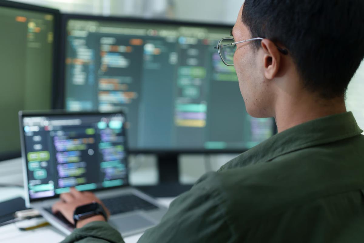 An engineer in glasses and an olive shirt works on a laptop in front of large monitors showing code, illustrating a developer testing Claude Opus 5.5 and GPT-6 Astra on agentic coding and long-running engineering tasks
