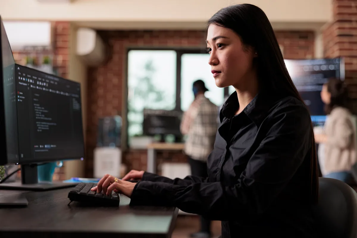 Forward-deployed engineer writing code at her desk while colleagues work in the background, as an embedded builder FDE would alongside a customer’s team.