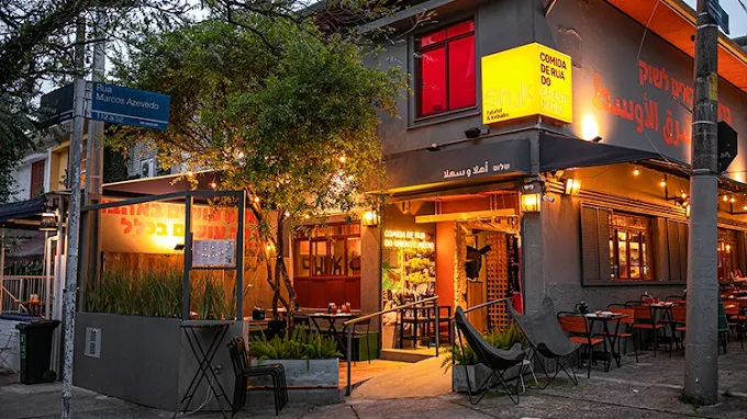 Fachada iluminada do Shuk em Pinheiros de comida de rua à noite em esquina com mesas externas e placas em português e hebraico.