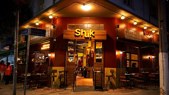 Fachada do restaurante Shuk Esfihas iluminado à noite com mesas e cadeiras do lado externo.