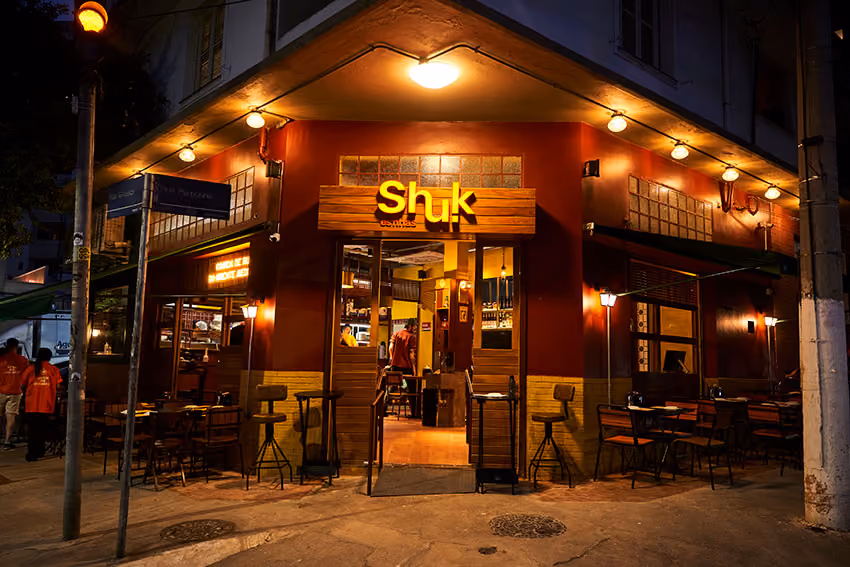 Fachada do restaurante Shuk Esfihas iluminado à noite com mesas e cadeiras do lado externo.