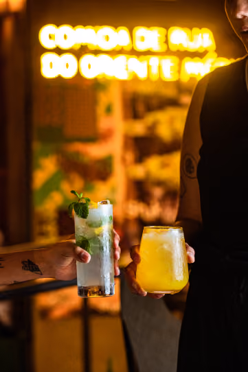 Dois drinques, um com hortelã e outro amarelo com gelo, erguidos diante do neon comida de rua do Oriente Médio