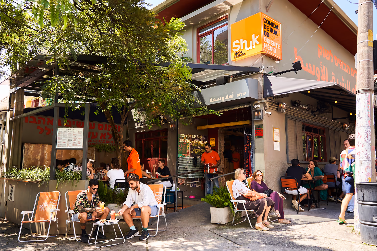 Calçada do Shuk Falafel & Kebab num dia de sol, com clientes nas cadeiras de lona e a equipe de camiseta laranja