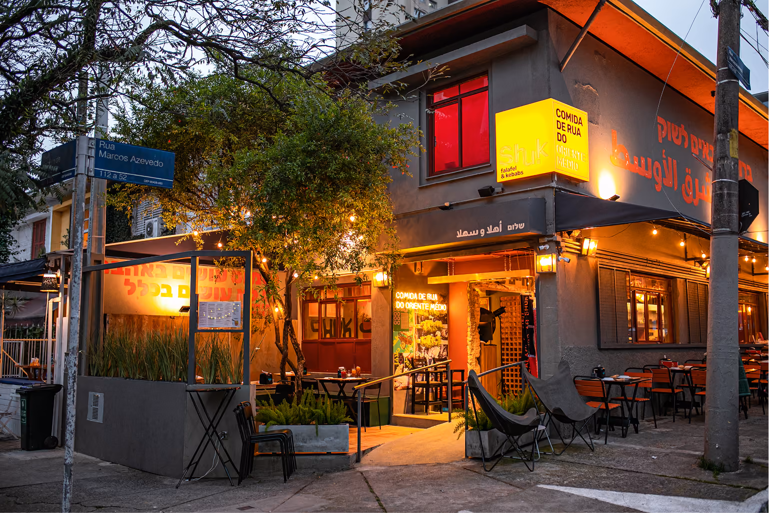Entrada do Shuk Falafel & Kebab ao anoitecer, com a árvore da esquina, o letreiro aceso e cadeiras de lona na calçada