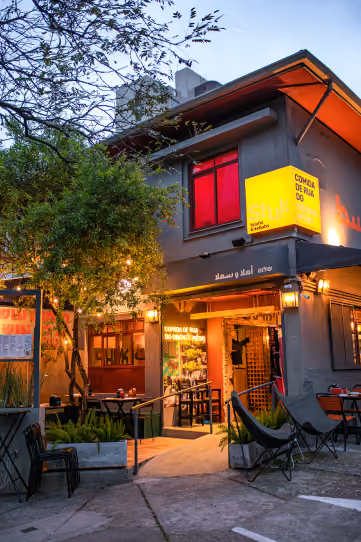 Entrada do Shuk Falafel & Kebab ao anoitecer, com a árvore da esquina, o letreiro aceso e cadeiras de lona na calçada