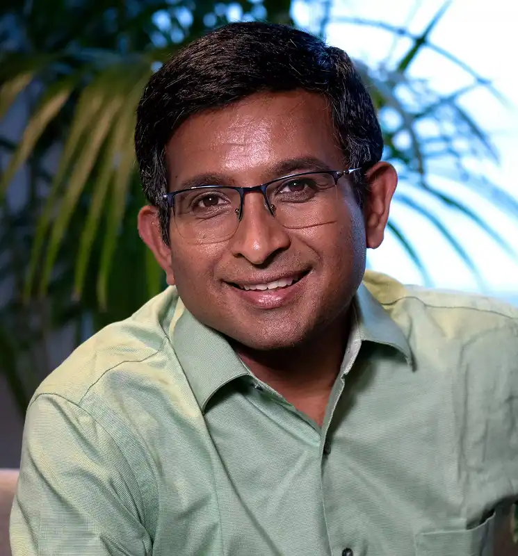 Smiling man with short black hair and glasses wearing a light green shirt with plants in the background.