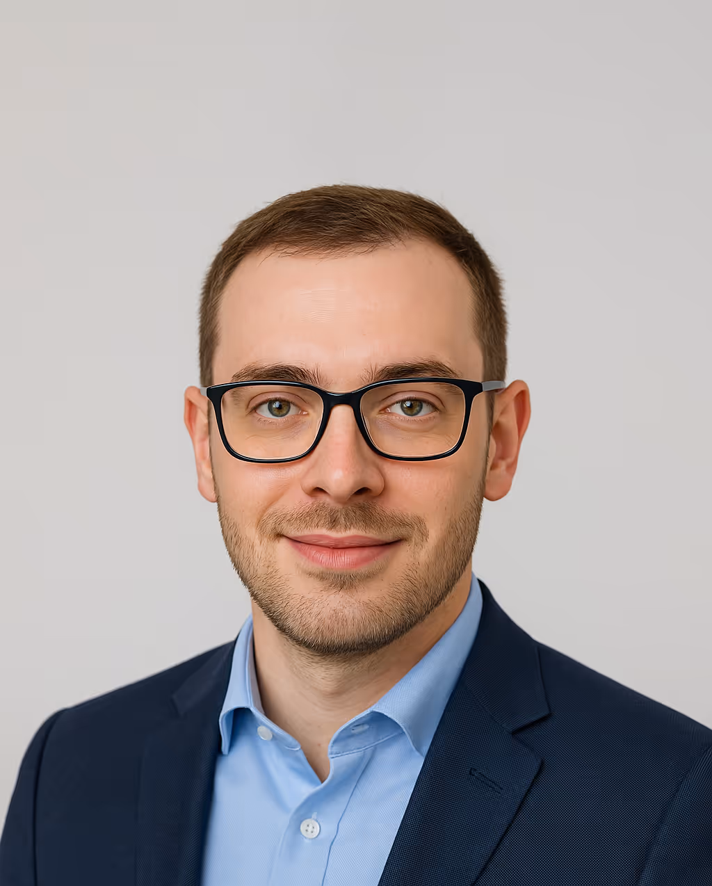 Smiling man with short brown hair, glasses, blue shirt, and navy blazer against a plain background.