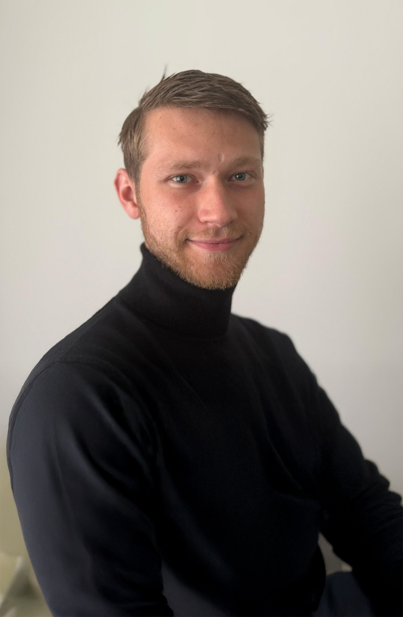 Young man with light beard and short brown hair wearing a black turtleneck sweater, smiling gently against a plain light background.
