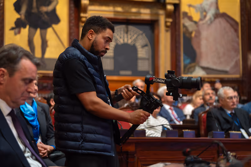 A man operating a Ronin camera stabilizer filming a seated audience in a formal room with classical paintings.