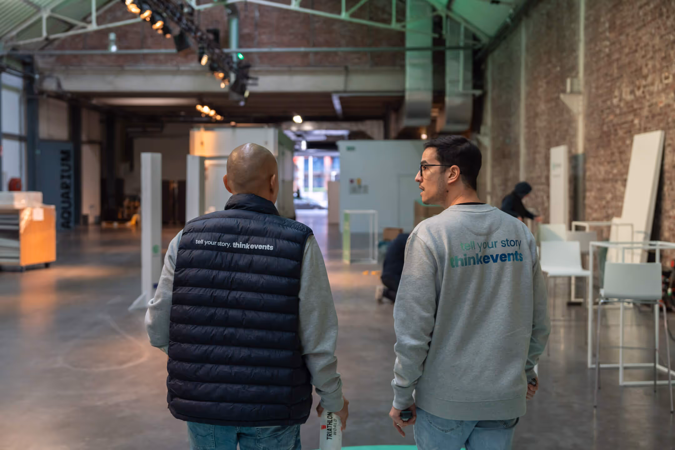 Two men wearing jackets with thinkevents logos on the back, standing and facing away inside a large industrial-style room with exposed brick walls and high ceilings.