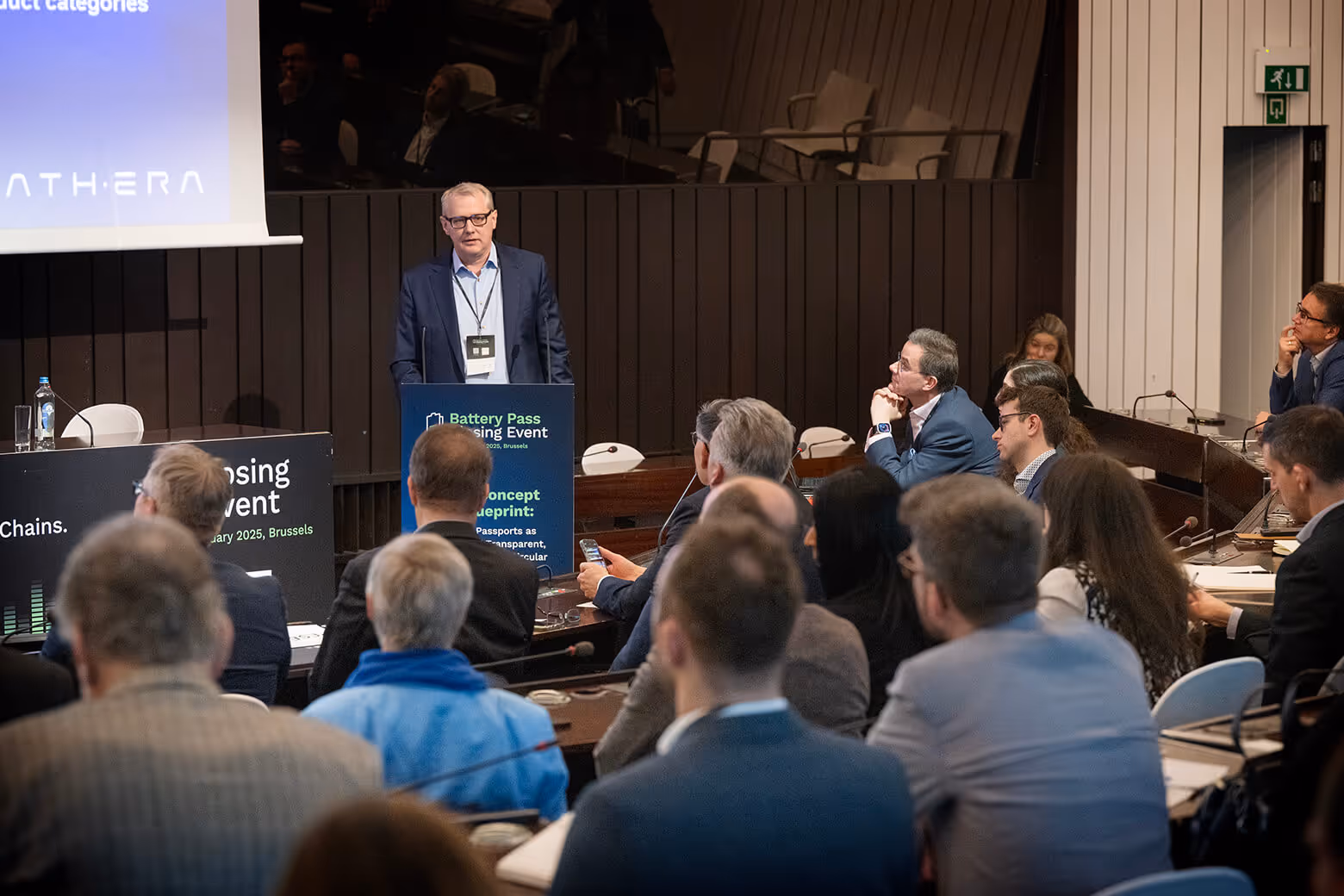 Man in a suit and glasses speaking at a podium during the Battery Pass Closing Event with an audience seated and listening attentively.