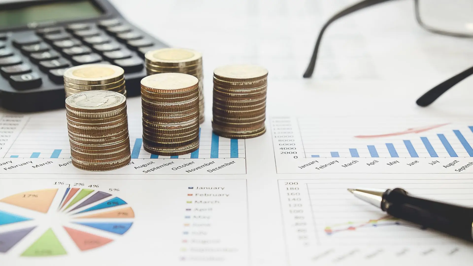 A close-up shows four stacks of gold and silver coins placed directly on printed financial reports. The documents display blue bar charts, a colorful pie chart, and tables. In the slightly blurred background are a black calculator and a pair of glasses, while an elegant pen lies in the right foreground. The image symbolizes the topics of financial planning, controlling, data-driven business management, and the digitalization of the finance function.