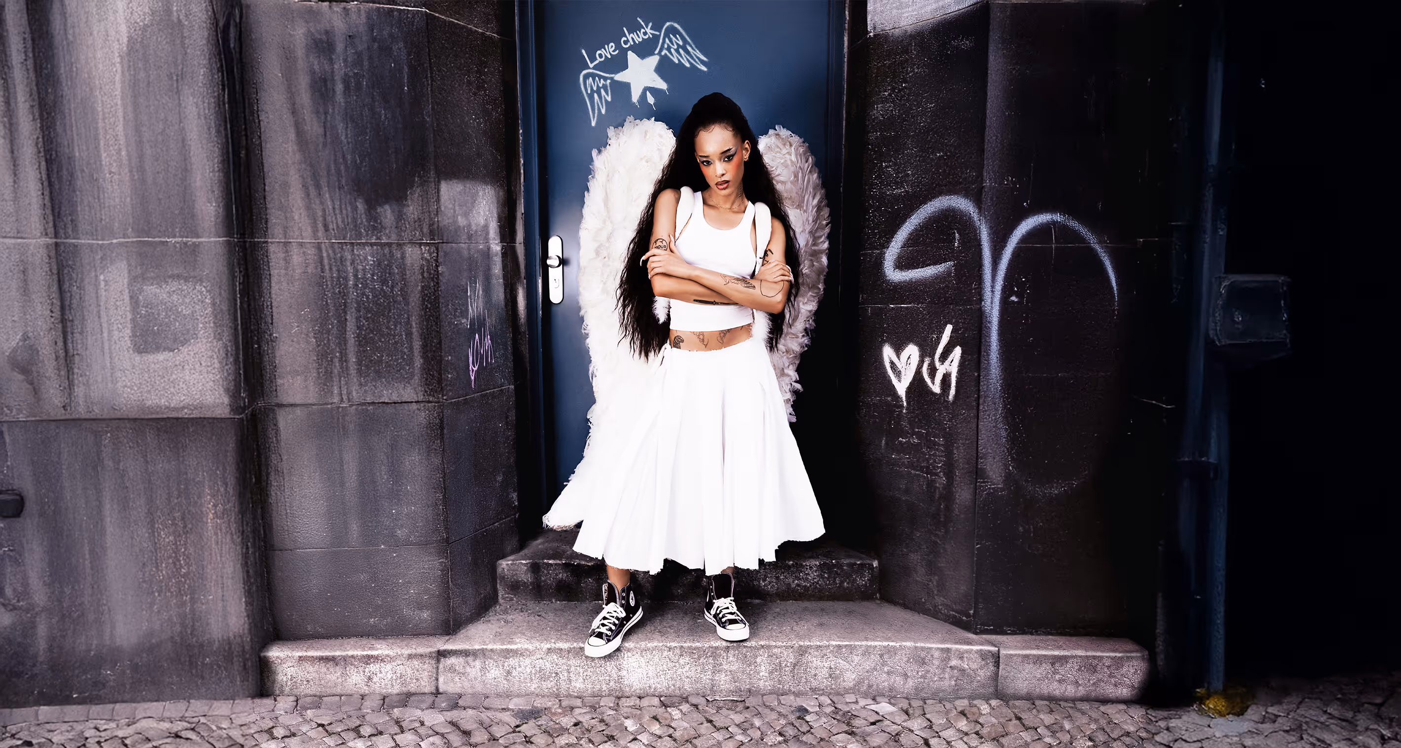 A model with tattoos standing in front of a building with a blue door and she is wearing angel wings.