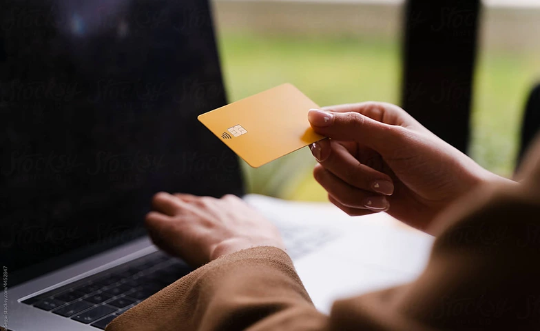 Person holding a yellow contactless credit card near a laptop keyboard.