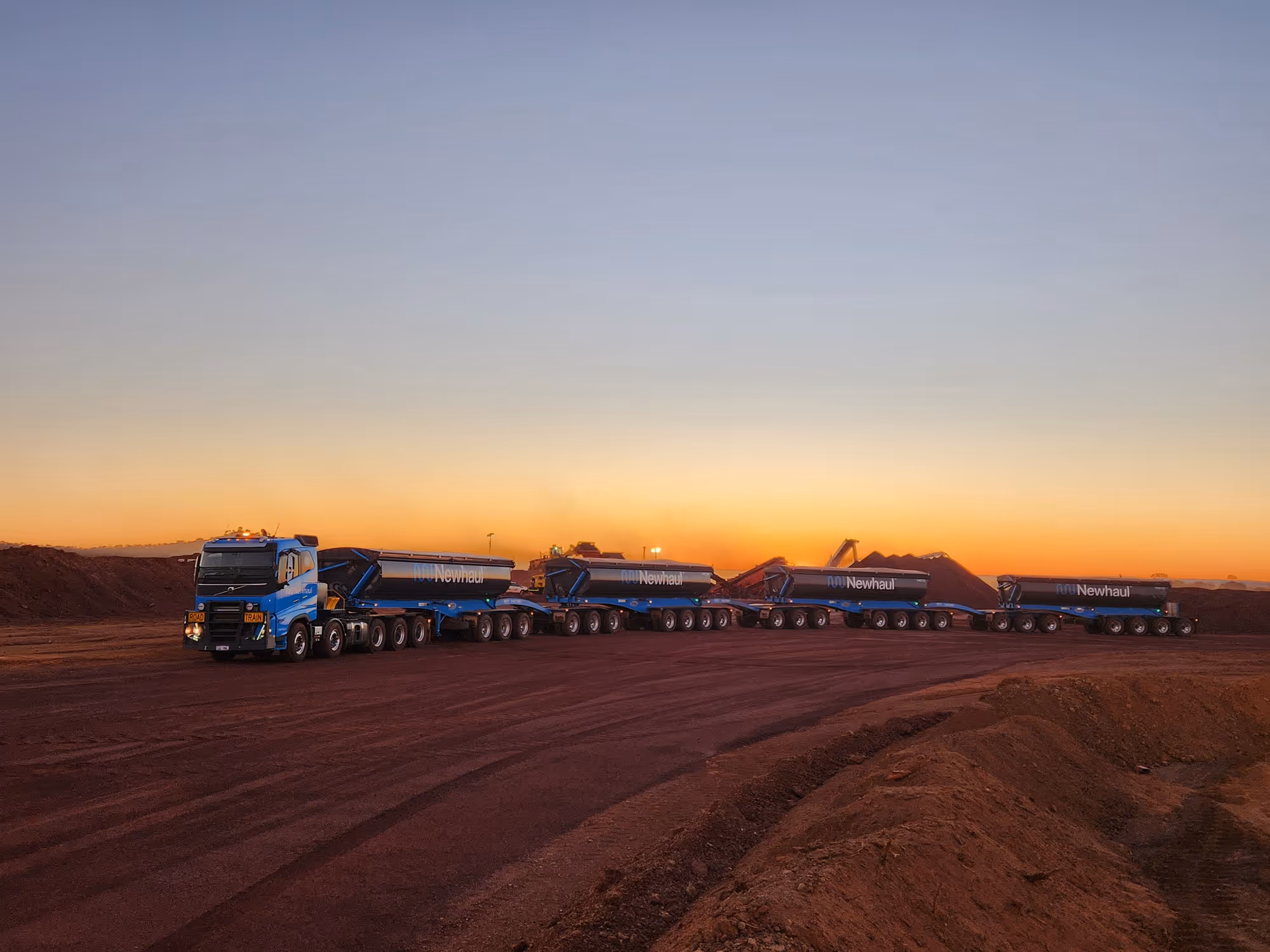Long articulated road train truck with four blue Newhaul trailers on a red dirt road at sunset.