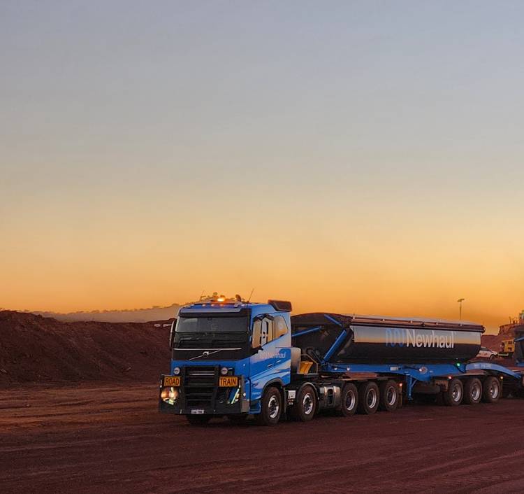 Blue Road Train truck hauling cargo on a dirt road during sunset with orange sky.