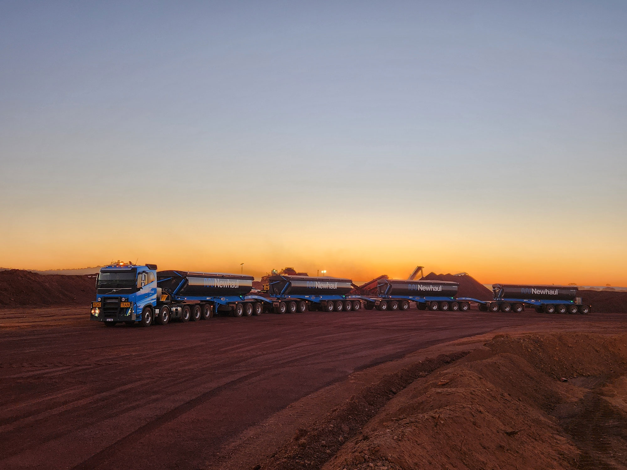 Long blue road train truck with four trailers on a dirt road at sunset in a mining site.