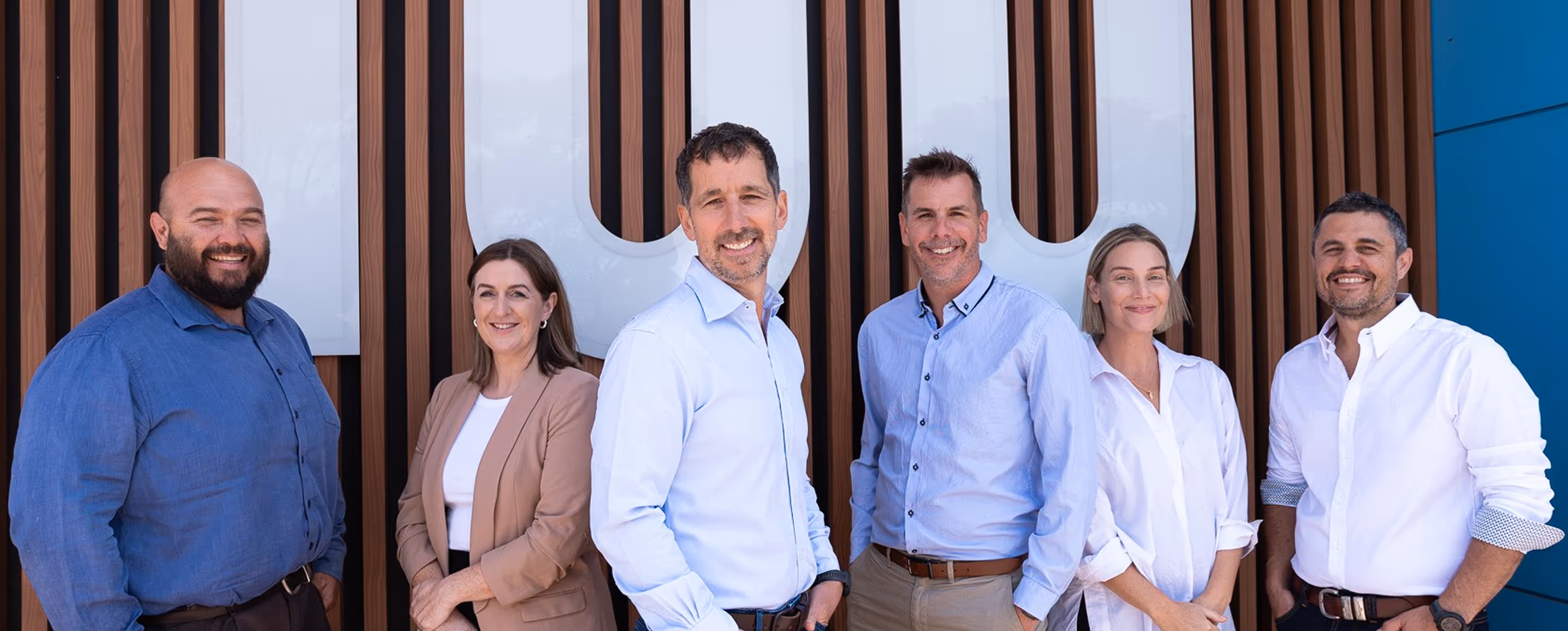 Group of six smiling professionals standing outdoors in front of a modern wall with wood paneling and large white letters.
