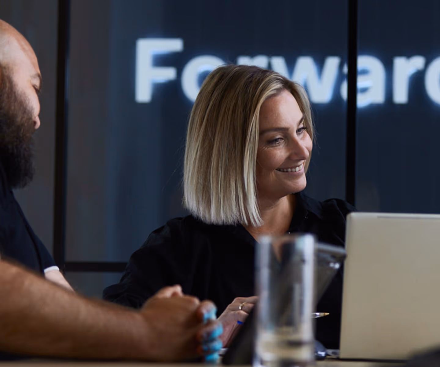 Smiling woman with blonde hair looking at a laptop next to a man with a beard in a meeting room.