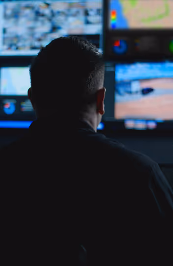 Man viewed from behind monitoring multiple digital screens displaying maps and data in a dark control room.