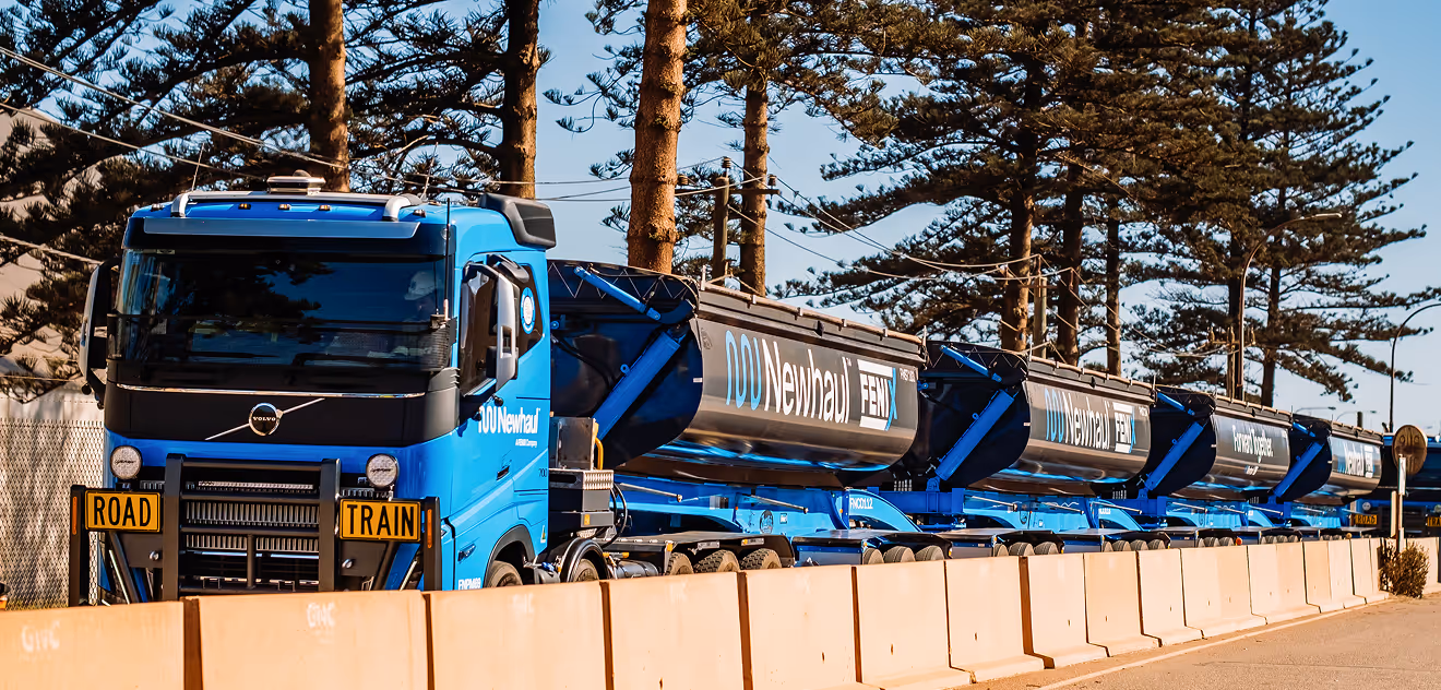 Blue multi-trailer road train truck parked alongside concrete barriers and tall pine trees.