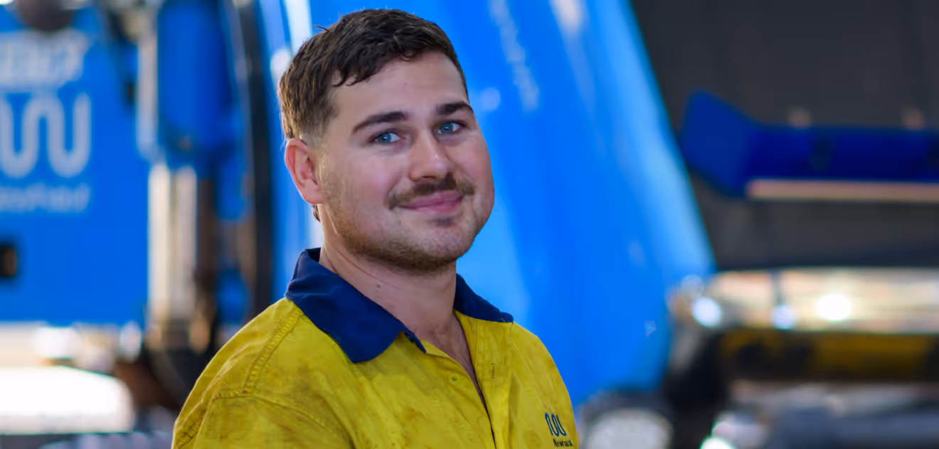 Smiling man wearing a yellow and navy work shirt with a blurred industrial background.