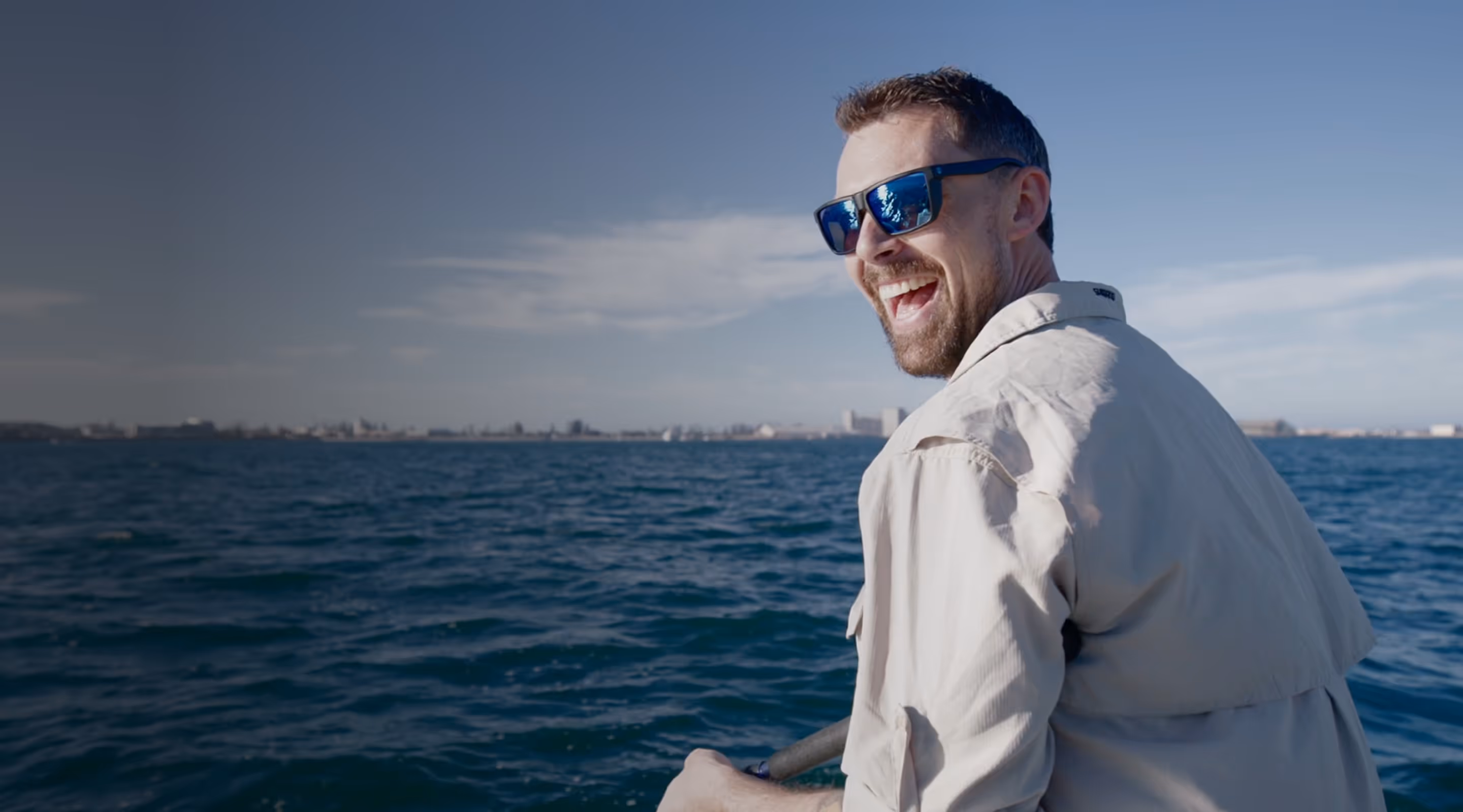 Smiling man wearing sunglasses and a beige jacket looks back while standing on a boat over a blue ocean with a city skyline in the distance under a clear sky.